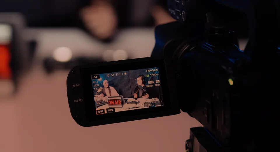 Behind-the-scenes view of a video camera recording two women in a studio during a broadcast, with the live camera screen showing them seated at a desk with microphones and an "ON AIR" sign.