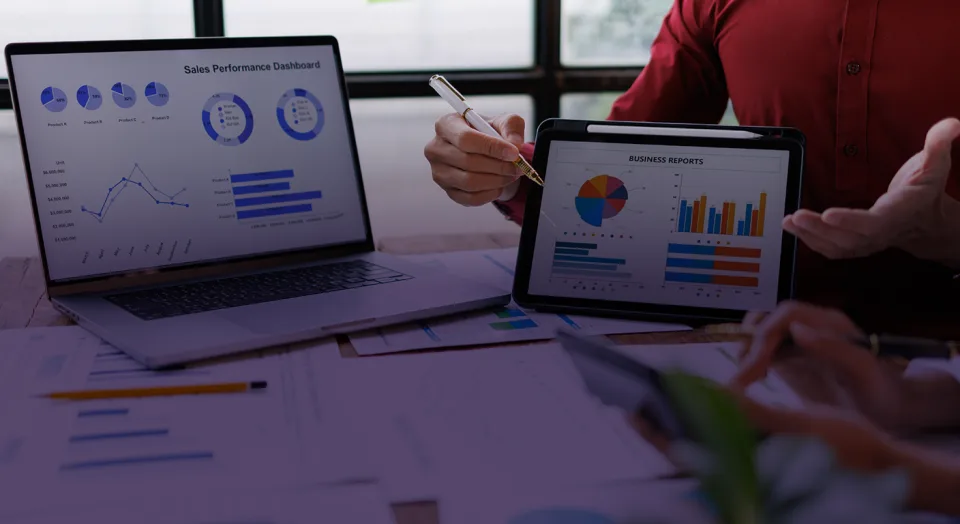 A business meeting setting with two people reviewing sales analytics and financial performance charts on a laptop, a tablet, and printed reports at a wooden desk.