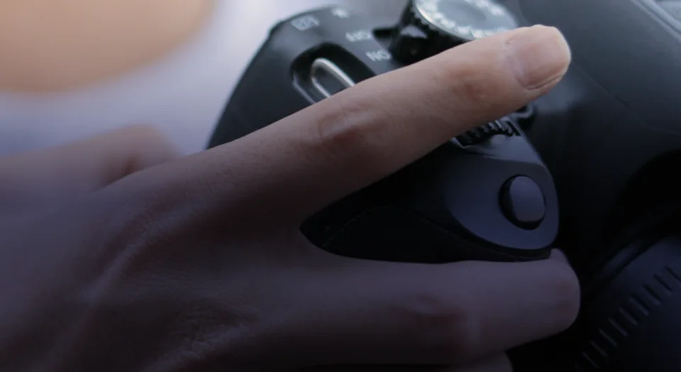 Macro shot of a hand gripping a black DSLR camera body, highlighting the finger resting near the shutter button, control dial, and power switch.